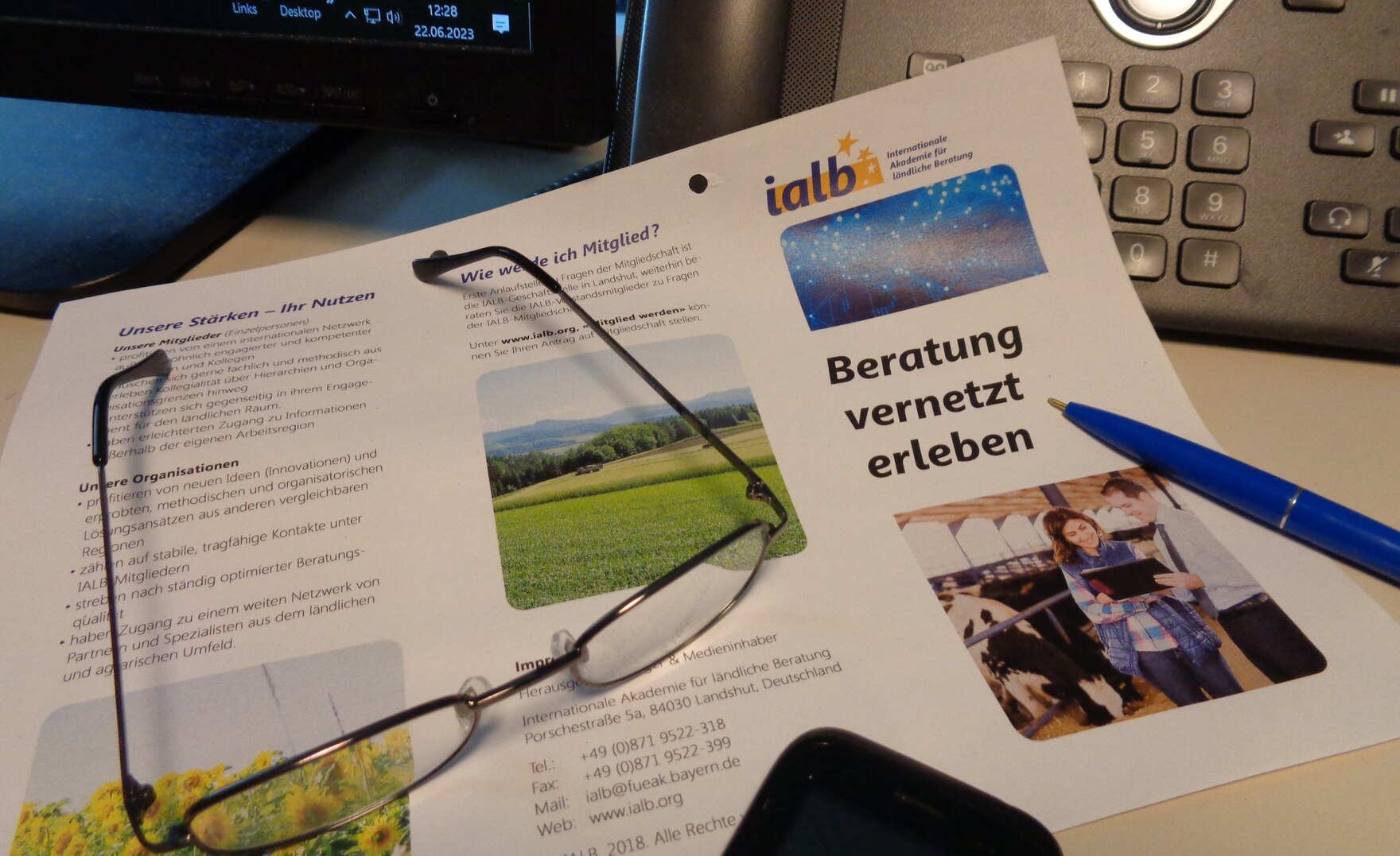 Flyer IALB, Brille liegt auf Flyer, Stift, Ausschnitt von Teleon und Monitor im Hintergrund
